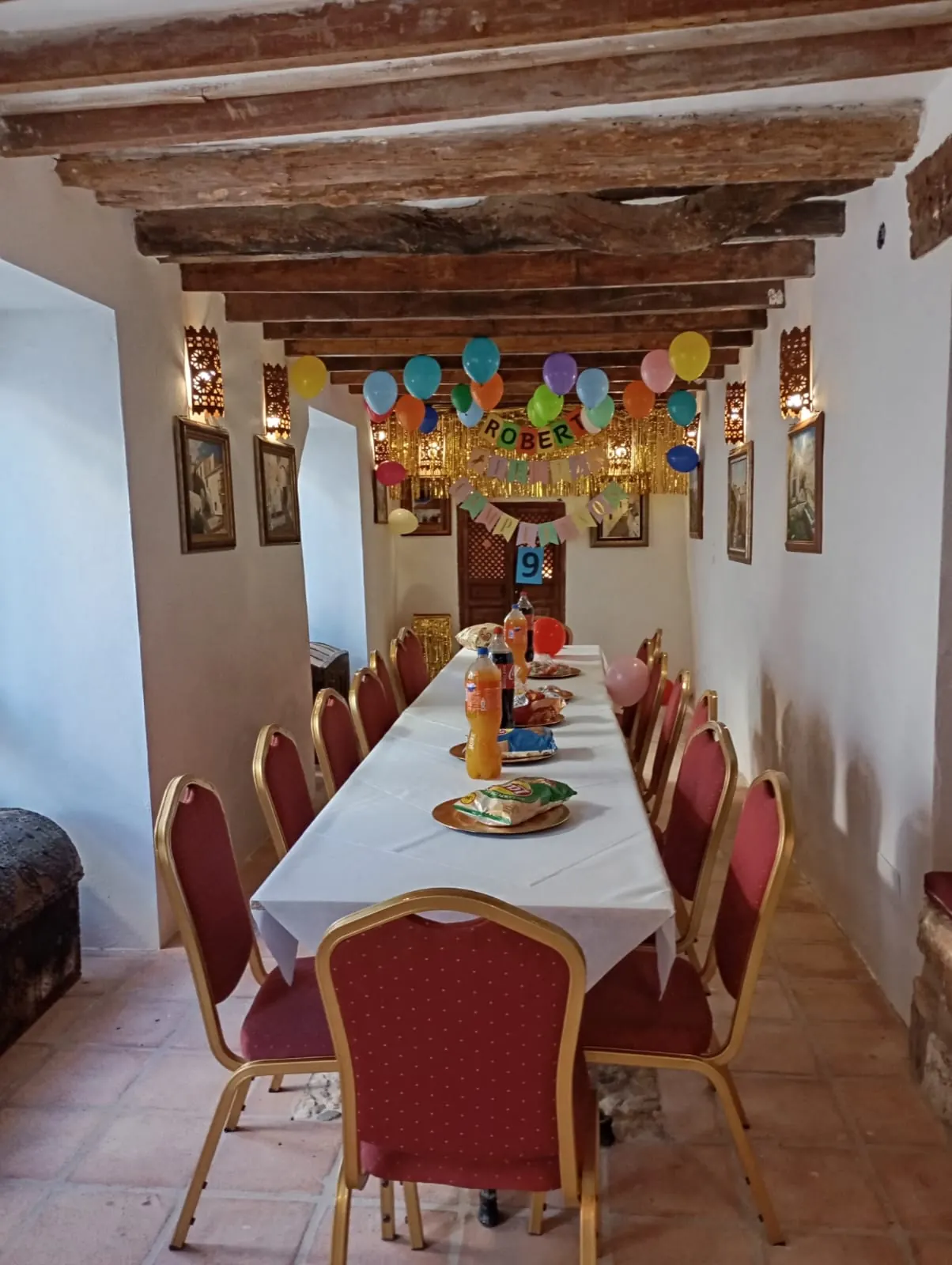 Mesa larga con sillas granates preparada para un cumpleaños, con globos en el techo
