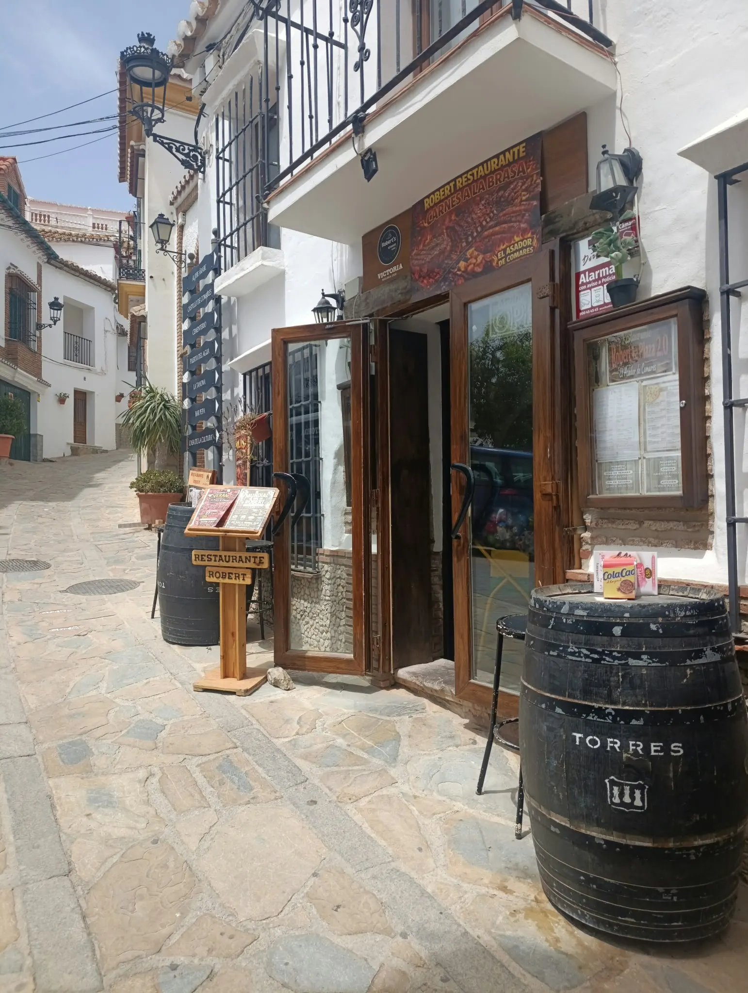 Puerta de madera de Restaurante Robert en una calle blanca de Comares, con un barril a la entrada